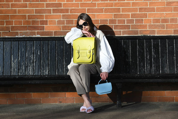 woman sat on a bench holding handmade leather backpack in yellow and Aura handbag in baby blue