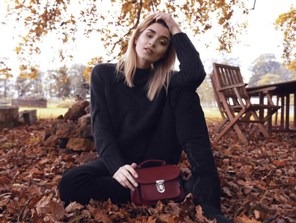 woman sat on leafy ground in autumnal setting with luna handbag in red placed in front of her