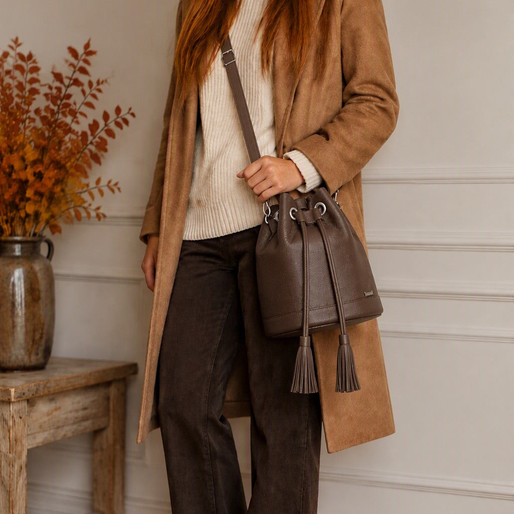 A person wearing a long tan coat, cream ribbed sweater, and dark brown pants carries a dark brown leather bucket bag with a drawstring closure and tassel details against a white paneled wall.