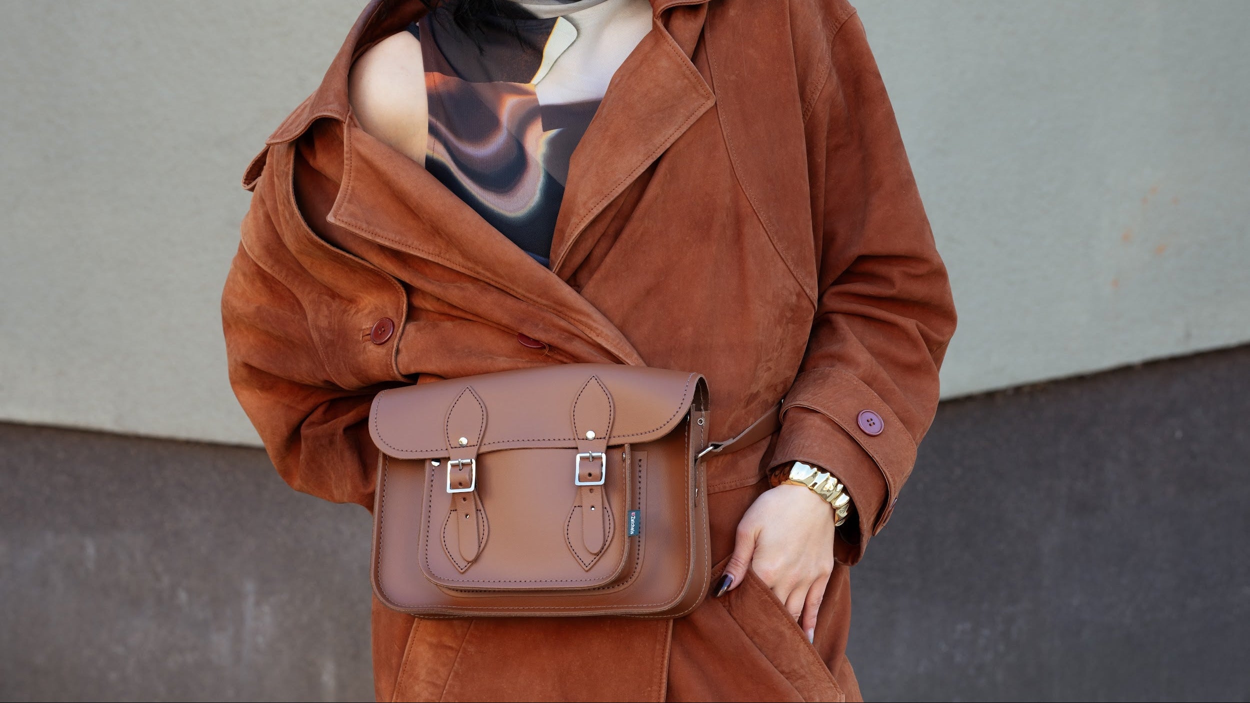 A woman wearing a brown suede trench coat and a patterned top, carrying a matching brown leather Satchel-style crossbody bag against a gray wall.