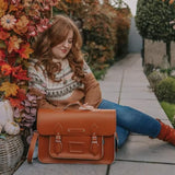 Our model, Emily, is sitting on the ground next to autumnal leaves, with a 16'' Burnt orange Zatchels satchel on the ground next to her.