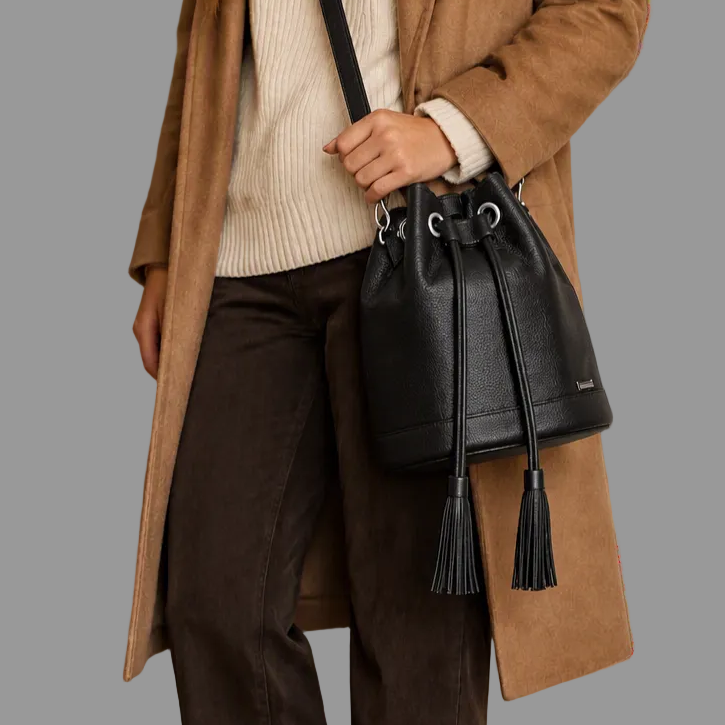 A person wearing a camel coat, cream ribbed sweater, and dark brown pants carries a black leather bucket bag with a drawstring and tassel against a white wall.