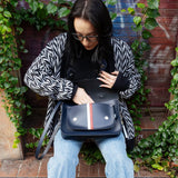 Woman holding a navy satchel handbag with a red and white stripe against a leafy background