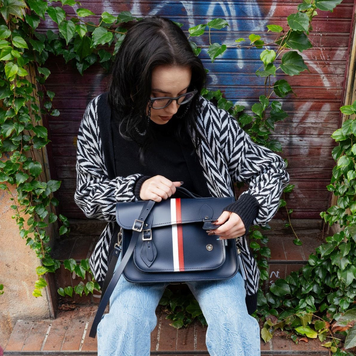 Person holding a navy satchel with red and white stripe against a graffiti-covered wall.