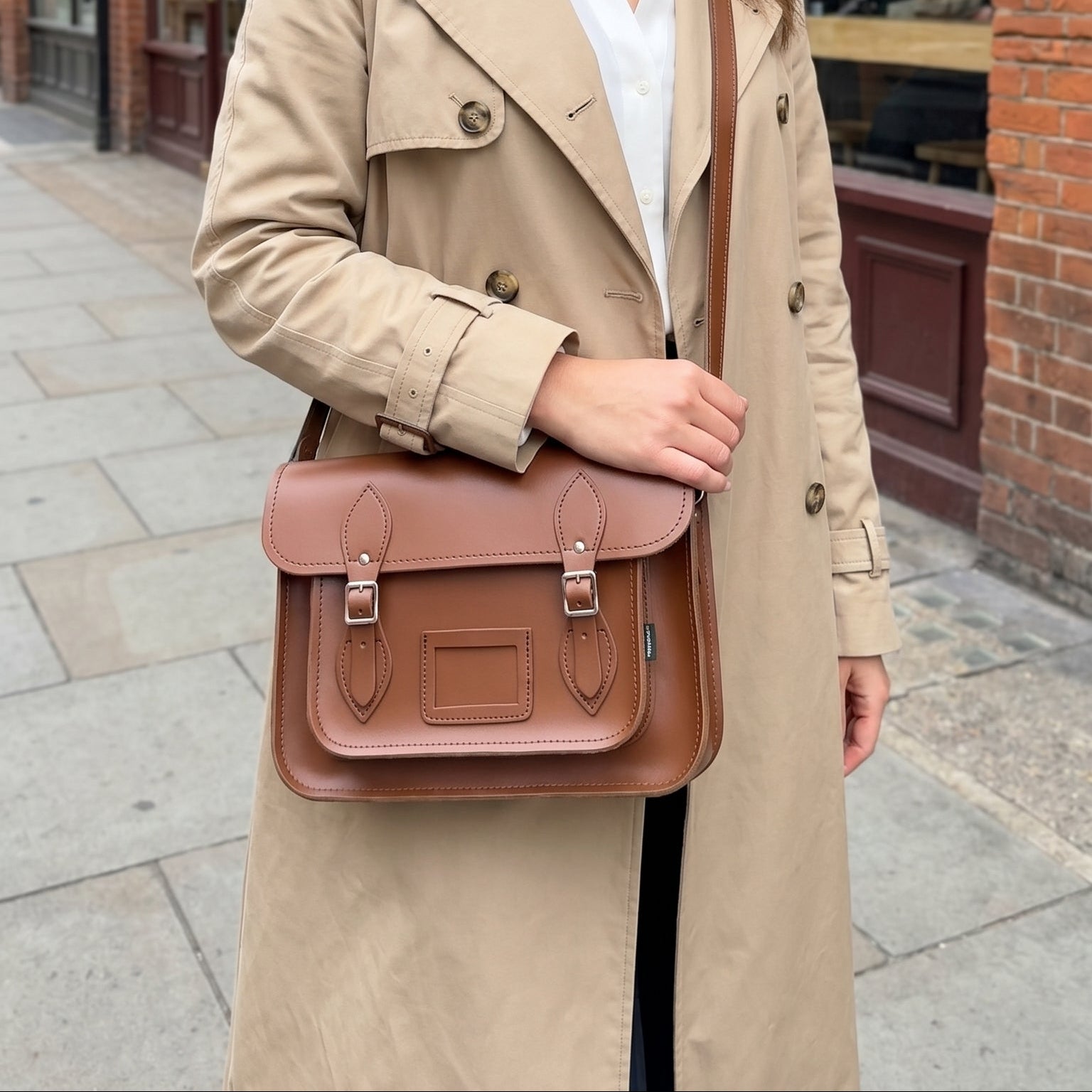 Woman in a trench coat holding a brown satchel bag on a city street.
