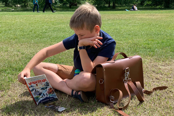 boy sat reading a book on a grass lawn with Zatchels handmade leather satchel backpack in brown placed next to him