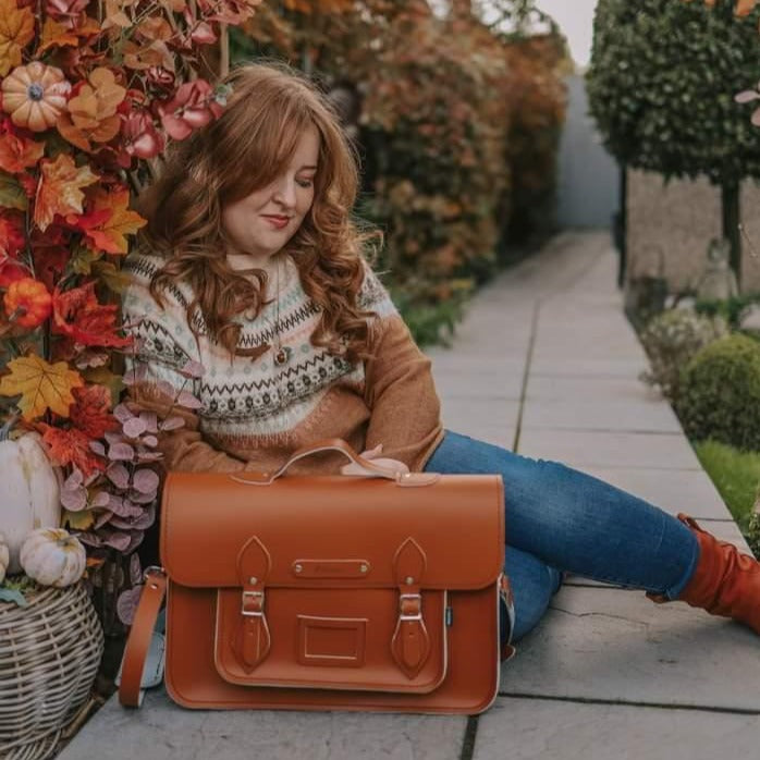 Our model, Emily, is sitting on the ground next to autumnal leaves, with a 16'' Burnt orange Zatchels satchel on the ground next to her. 