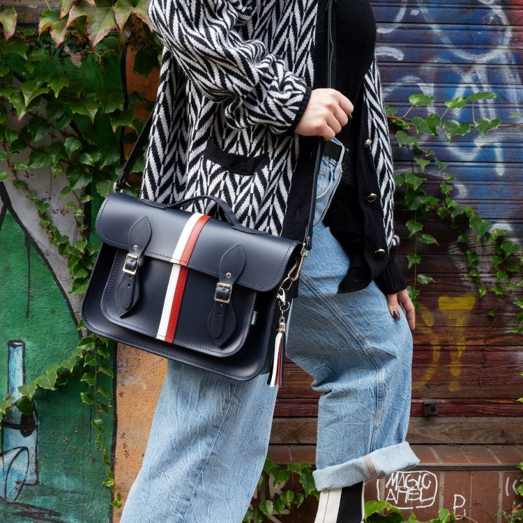 Person holding a navy satchel with red and white strripe against a graffiti-covered wall.