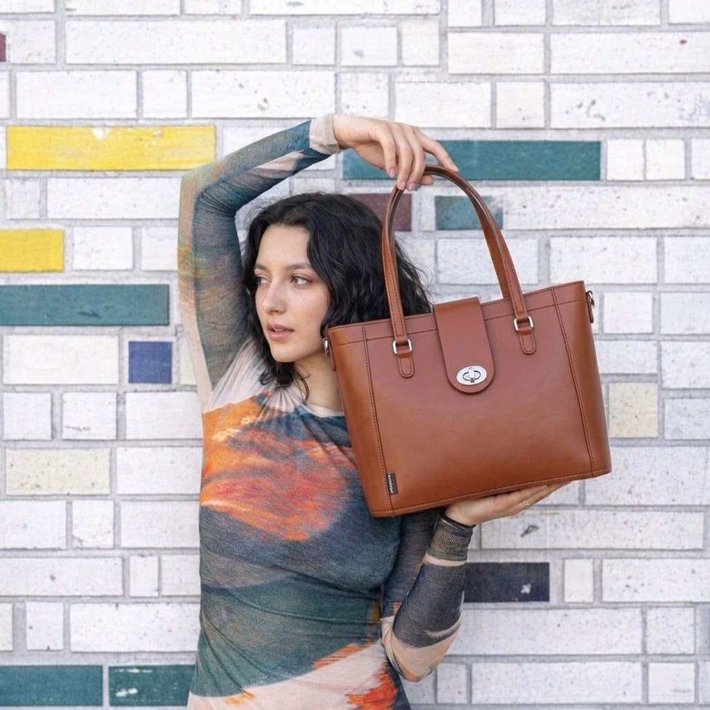 A woman holding a brown leather tote bag in front of a white brick wall with colorful painted accents.