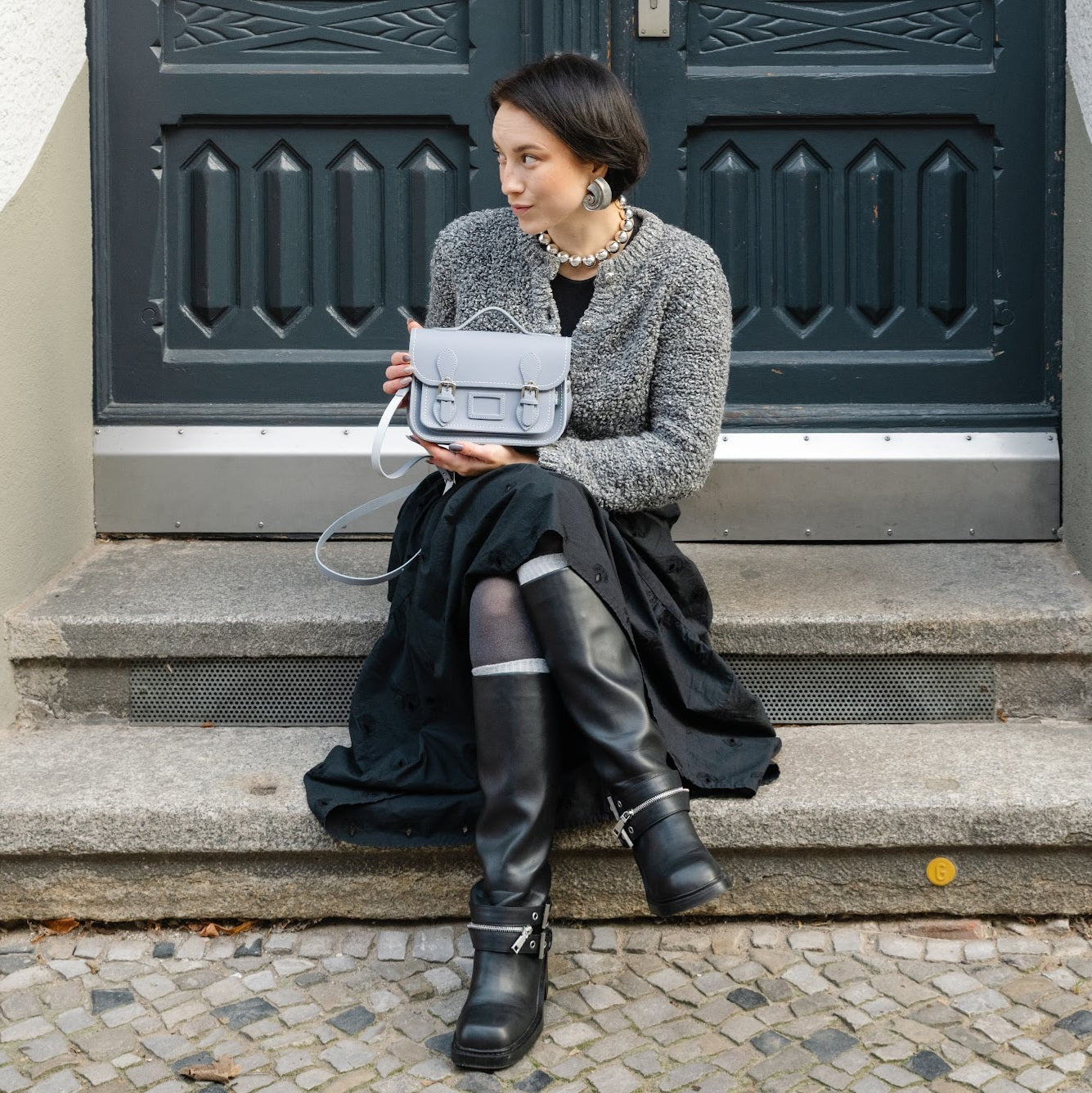 Woman sitting on steps holding a gray handbag in front of a dark green door.