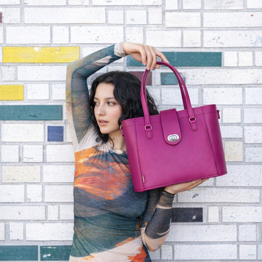 A woman wearing a colorful long-sleeve top holds up a bright magenta leather tote bag in front of a white brick wall with colored accent blocks.