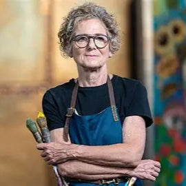 Artist with a blue apron holding paintbrushes in front of a colorful abstract painting.